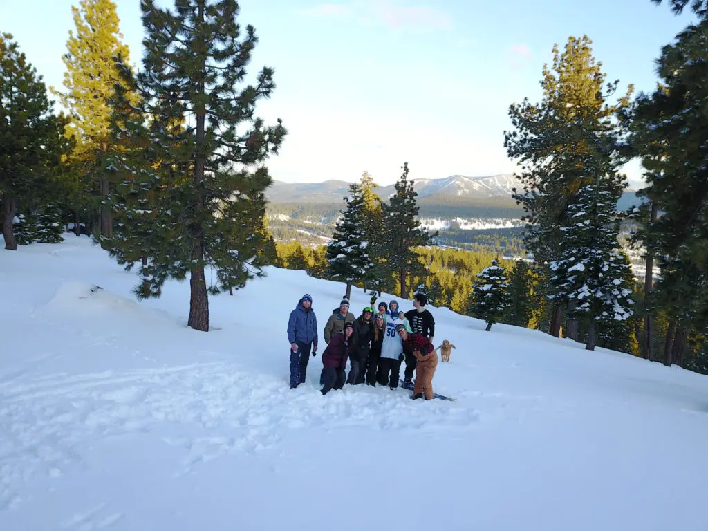 Nio personer står i snön i sluttande landskap med träd runtomkring och utsikt över berg.