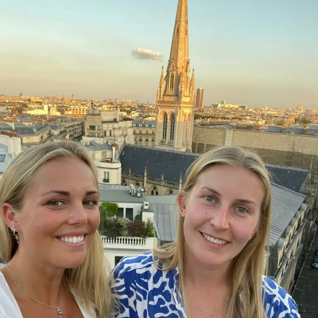 Två blonda kvinnor ler mot kameran. I bakgrunden stadsmiljö och ett gammalt elegant torn.
