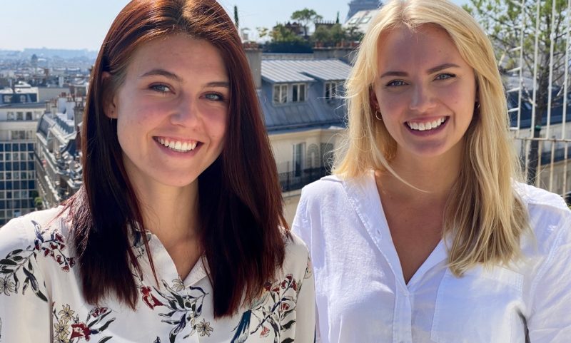 Sara och Hanna utomhus med stad i bakgrunden. Sara har blont långt hår, vit skjorta. Hanna har brunt långt hår och en vit skjorta med blommigt mönster.