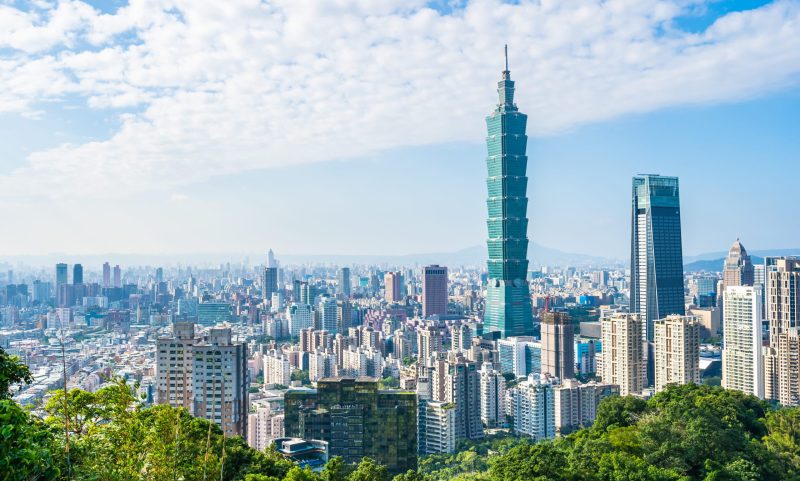 Stadsvy över Taipei City. Två höghus sticker upp ovanför övriga höghus en ljus sommardag med grönska i förgrunden.